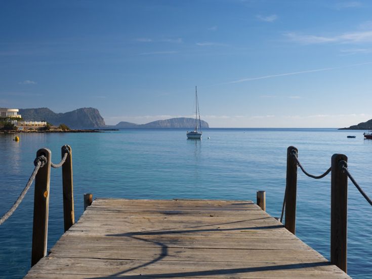 Holzsteg in Es Canar auf Ibiza mit Blick auf das ruhige Meer und ein Segelboot am Horizont