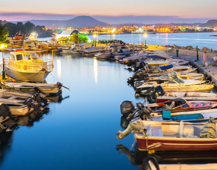 Kleiner Hafen mit mehreren Booten am Ufer, beleuchtete Stadt und Berge im Hintergrund bei Abenddämmerung.