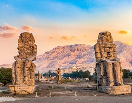 Zwei massive, verwitterte Kolossalstatuen der Memnon-Kolosse stehen vor einer Berglandschaft bei Sonnenuntergang