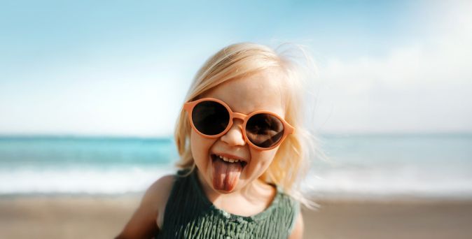Ein kleines Kind mit großer, orangefarbener Sonnenbrille steht am Strand vor dem Meer, bei hellem Sonnenlicht und leichtem Wind und streckt dabei die Zunge heraus, während der Sandstrand und die Meereswellen im Hintergrund sichtbar sind.