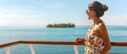 Frau steht an der Reling eines Kreuzfahrtschiffs und blickt auf das ruhige, türkisfarbene Meer mit einer kleinen Insel am Horizont