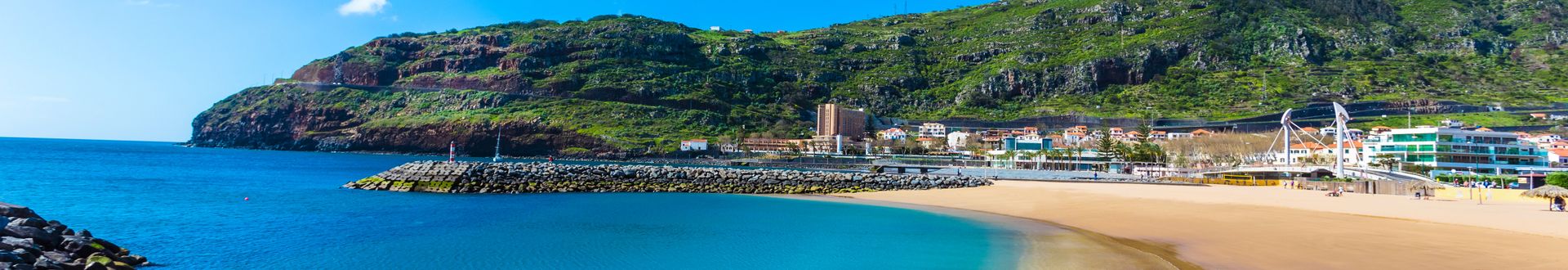Sandstrand Praia de Machico auf Madeira mit geschützter Badebucht, türkisblauem Wasser, Wellenbrechern und grünen Berghängen im Hintergrund