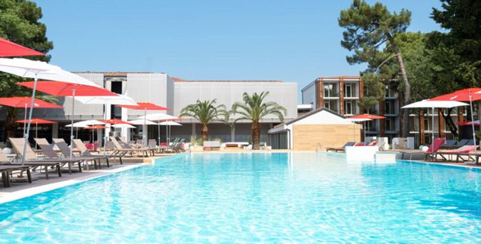 Großer Außenpool eines Hotels mit türkisfarbenem Wasser, umgeben von Liegestühlen mit roten Sonnenschirmen, Palmen und modernen Hotelgebäuden unter blauem Himmel.