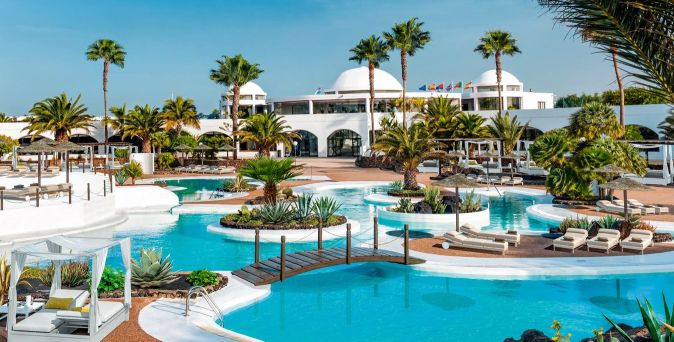 Ein Resort mit Pool, umgeben von Palmen und Sonnenliegen. Im Hintergrund ein weißes Gebäude mit Kuppeldach und Flaggen.