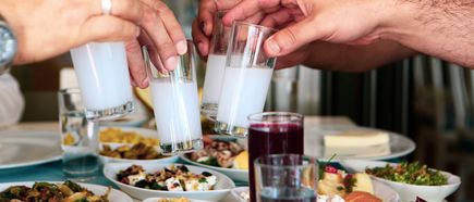 Mehrere Hände stoßen mit Gläsern Ouzo über einem gedeckten Tisch mit verschiedenen Speisen an
