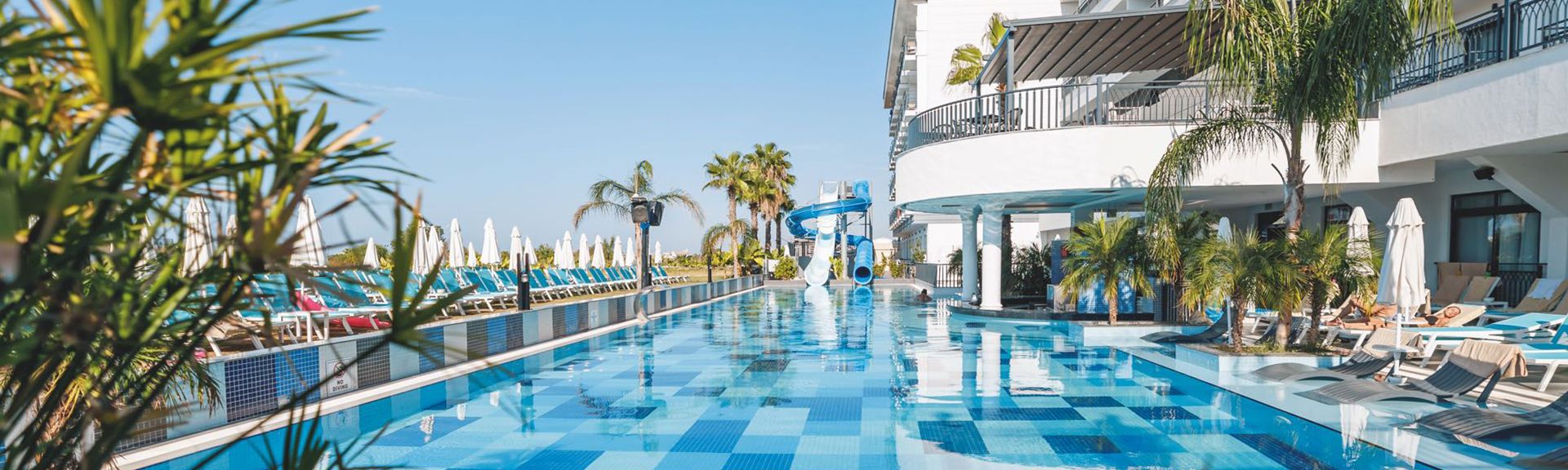 Großer Außenpool mit blau-weiß kariertem Boden, umgeben von Liegestühlen und Sonnenschirmen, angrenzend an ein mehrstöckiges Hotelgebäude mit Balkonen und Palmen im Hintergrund