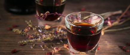 Nahaufnahme eines Glases mit dunklem Kräuterlikör, dekoriert mit getrockneten Kräutern auf einem Holztisch