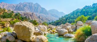 Felsige Landschaft der Wadi-Tiwi-Schlucht im Oman mit natürlichen Wasserbecken, Palmen und Gebirgszügen im Hintergrund