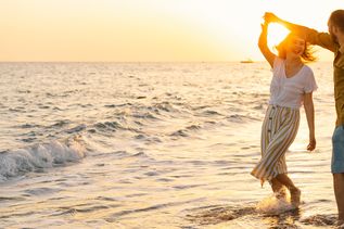 Ein Paar tanzt am Strand im flachen Wasser bei Sonnenuntergang, die Frau trägt ein weißes T-Shirt und gestreiften Rock, der Mann ein braunes Hemd und blaue Shorts.