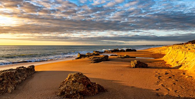 Strand mit Felsen und goldfarbenem Sand bei Sonnenuntergang unter bewölktem Himmel