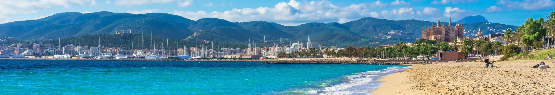 Panoramaaufnahme des Strandes von Playa de Palma auf Mallorca mit türkisblauem Meer, feinem Sand, Yachthafen und der Kathedrale La Seu im Hintergrund vor einer grünen Bergkulisse