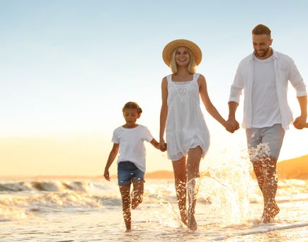 Familie mit zwei Kindern läuft am Strand bei Sonnenuntergang, alle tragen helle Kleidung