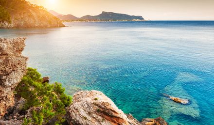 Felsige Küste bei Kemer in der Türkei im warmen Sonnenuntergang, mit türkisblauem Meer, klarem Wasser und bewaldeten Bergen im Hintergrund