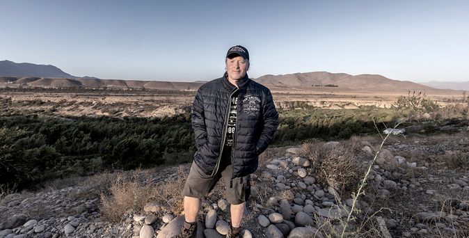 Joey Kelly mit schwarzer Jacke, Shorts und Basecap steht auf felsigem Boden vor einer weiten, kargen Landschaft mit Bergen im Hintergrund