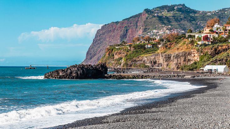 Kieselstrand Praia Formosa auf Madeira mit Atlantikwellen, dunklen Felsformationen, Steilküste und Häusern auf den terrassierten Berghängen im Hintergrund