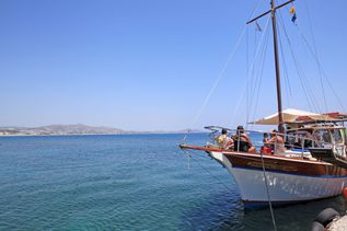 Mehrere Personen sitzen auf einem hölzernen Segelboot, das an einem Steg im Meer vor einer Küstenlandschaft festgemacht ist.