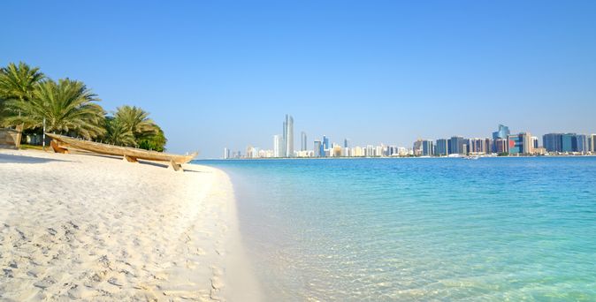 Strand mit Palmen und Booten, Blick auf die Skyline von Abu Dhabi am klaren Himmel