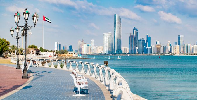 Skyline mit modernen Wolkenkratzern, Sitzbänken entlang der Uferpromenade und einer Flagge der Vereinigten Arabischen Emirate an einer Laterne