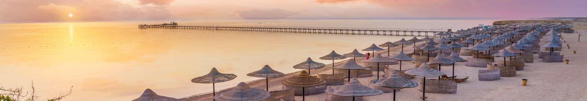 Ruhiger Strand in Marsa Alam bei Sonnenaufgang mit Reihen von Strohsonnenschirmen, leeren Liegen und einem langen Steg über dem goldschimmernden Meer