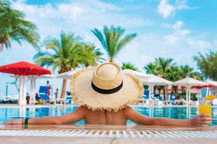Person mit Strohhut lehnt an der Kante eines Pools und hält ein Getränk, während Palmen, Sonnenschirme und Liegen im Hintergrund zu sehen sind