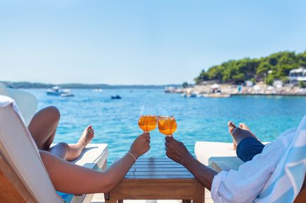 Zwei Personen stoßen mit orangenen Getränken in Gläsern an, während sie am Meer liegen, mit Blick auf Wasser und Himmel
