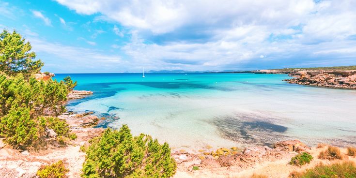 Cala Saona auf Formentera mit türkisfarbenem, flachem Wasser, rötlichen Felsen und grünem Küstenbewuchs unter leicht bewölktem Himmel