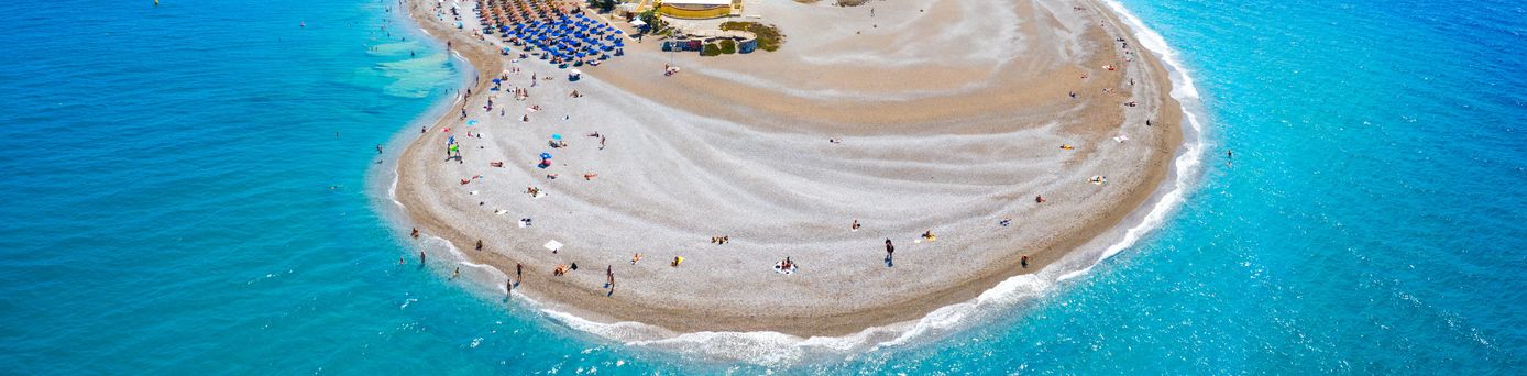 Luftaufnahme eines langen, gebogenen Sandstrands mit Sonnenschirmen und Gebäuden im Hintergrund an der Küste von Rhodos.