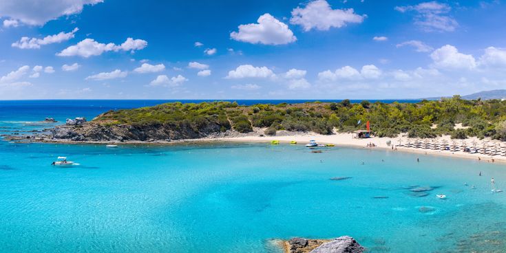 Blick auf die Glystra Beach auf Rhodos mit klarem türkisfarbenem Wasser, Sandstrand, Sonnenschirmen und Booten im Wasser.