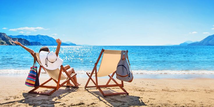 Person auf einem Strandstuhl an einem Sandstrand am Meer mit freiem Blick auf das glitzernde Wasser, einem leeren Strandstuhl daneben und Berglandschaften an der Küste im Hintergrund