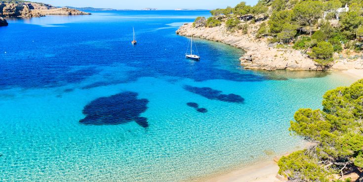 Bucht Cala Salada auf Ibiza mit türkisblauem Wasser, hellem Sandstrand, grünen Pinien und zwei Segelbooten im Meer