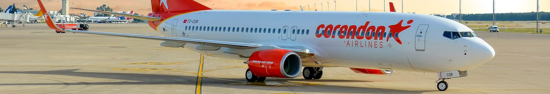 Boeing‑737‑800 der Corendon Airlines steht auf dem Vorfeld eines Flughafens, rote Triebwerke und Corendon‑Logo am Rumpf, Abfertigungsbereich und Rollwege im Hintergrund.