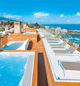 Dachterrasse mit mehreren Whirlpools und Liegestühlen, Blick auf Stadt, Berge und Meer unter blauem Himmel mit Wolken.