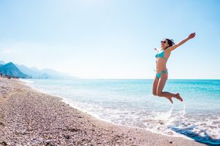 Frau im türkisfarbenen Bikini springt lachend am sonnigen Kiesstrand ins flache Meer, Berge im Hintergrund