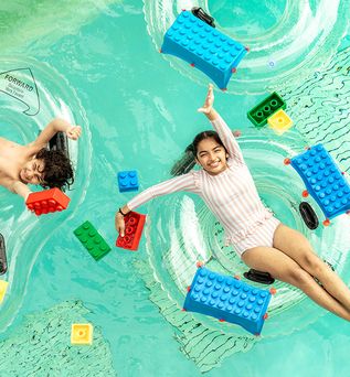 Drei Kinder liegen auf durchsichtigen Schwimmringen im Wasser, umgeben von großen, bunten LEGO-Steinen in Blau, Rot, Grün und Gelb.