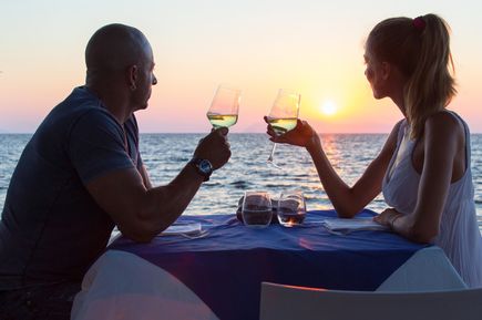 Ein Paar sitzt in einem Restaurant direkt am Meer bei Sonnenuntergang und halten Weingläser
