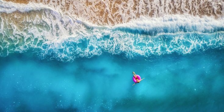 Luftaufnahme einer Frau in einem pinken Schwimmreifen, die entspannt im türkisblauen Meer nahe der Brandung treibt