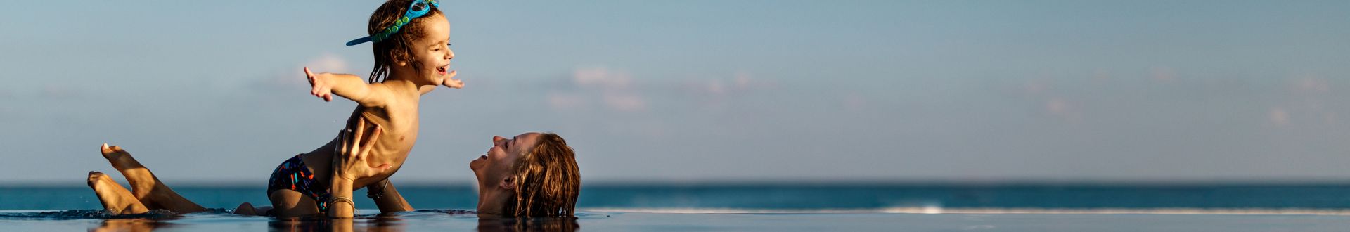 Erwachsene Person hebt ein Kind mit Tauchbrille in einem Pool hoch, während sich im Hintergrund Meer und Horizont erstrecken