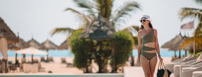 Frau im Badeanzug mit Strohhut geht am Pool entlang, im Hintergrund Palmen und Strandliegen