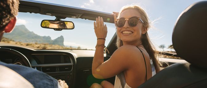 Junges Paar fährt in einem Cabrio bei Sonnenschein, die Frau mit Sonnenbrille lächelt fröhlich in die Kamera