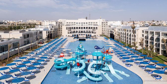 Großer Hotelpool mit mehreren blauen Wasserrutschen und Wasserspielgeräten umgeben von Reihen blauer Liegestühle auf beiden Seiten und weißen Hotelgebäuden im Hintergrund