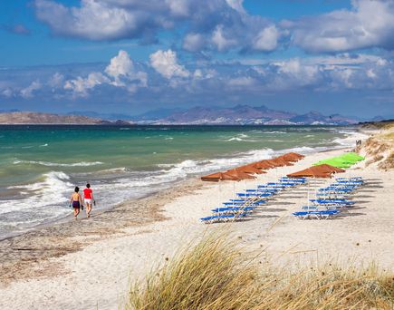Strand mit Reihen von blauen Liegestühlen und Sonnenschirmen sowie zwei Personen, die am Wasser entlanggehen, im Hintergrund Berge und bewölkter Himmel