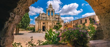 Blick durch einen steinernen Bogen auf das Kloster Arkadi mit barocker Fassade, blühenden Pflanzen und blauem Himmel mit Wolken.