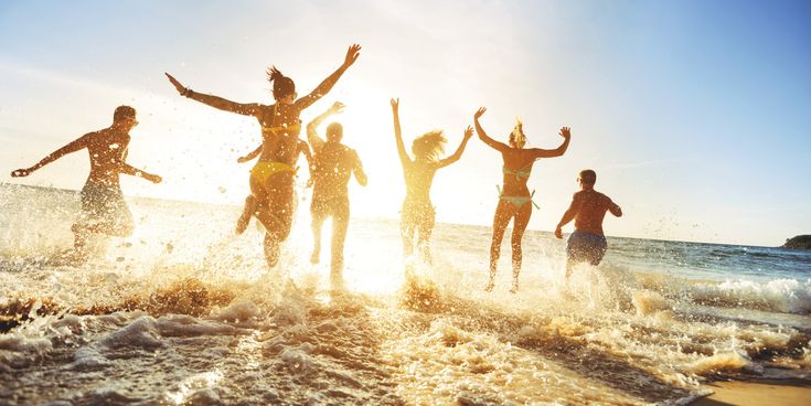 Gruppe junger Menschen rennt lachend und springend ins Meer, während im Sonnenlicht spritzende Wellen den Strand beleben