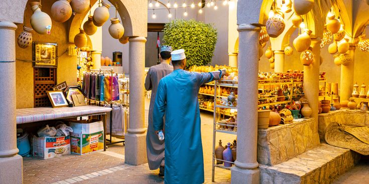 Zwei Männer in traditionellen Gewändern schlendern durch einen Markt mit Keramik, bunten Tüchern und Töpferwaren, die in warmem Licht ausgestellt sind