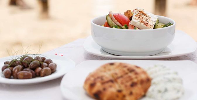 Weißer Teller mit griechischem Salat, daneben Teller mit Oliven und ein Teller mit Fladenbrot und Kräuterdip auf weißem Tischtuch.