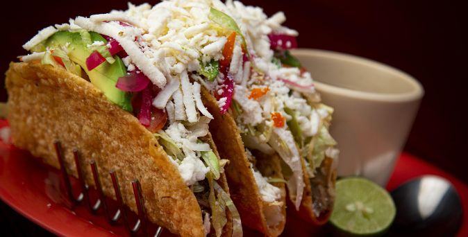 Drei knusprige Tacos gefüllt mit Avocado, roten Zwiebeln, Käse und Salat auf rotem Teller, daneben Limettenscheibe und Becher.