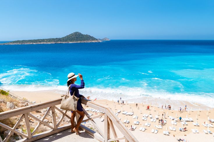 Person steht auf einer Holzterrasse mit Blick auf den Kaputas-Strand in der Türkei, türkisblaues Meer und weiße Sonnenliegen im Hintergrund
