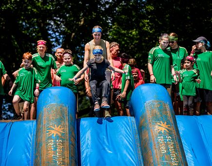 Kinder in grünen T-Shirts stehen auf einem blauen aufblasbaren Hindernis, ein Kind springt nach vorne
