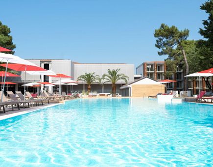 Großer Außenpool eines Hotels mit türkisfarbenem Wasser, umgeben von Liegestühlen mit roten Sonnenschirmen, Palmen und modernen Hotelgebäuden unter blauem Himmel.