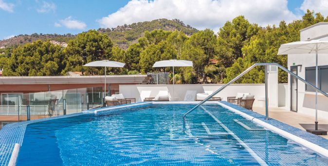 Ein Pool mit blauen Fliesen, umgeben von Liegen und Sonnenschirmen. Im Hintergrund sind grüne Bäume und Hügel sichtbar.